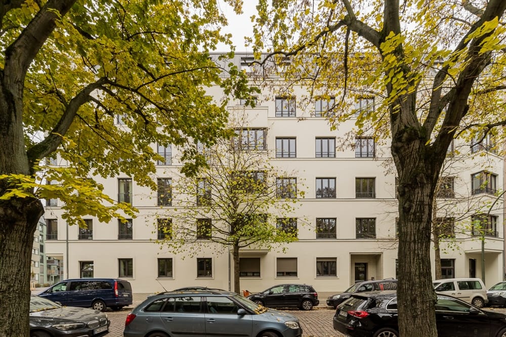 Widok z ulicy na pięciopiętrowy budynek wielorodzinny z białą elewacją otynkowaną i jesiennymi drzewami
