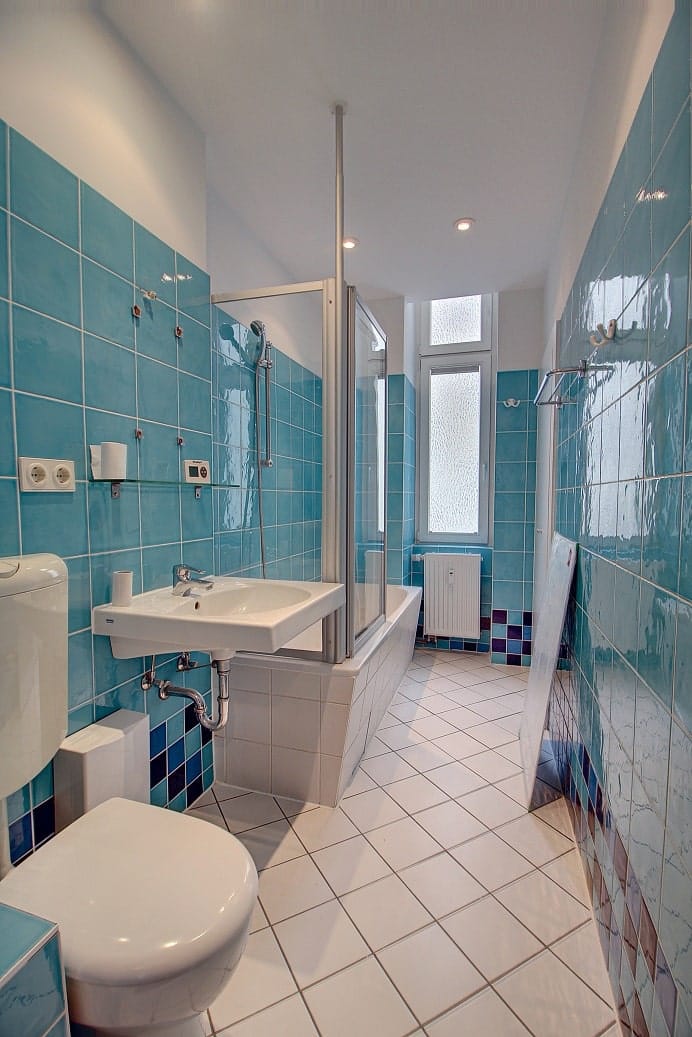 Interior view of a flat's bathroom with a bathtub and glass shower screen, as well as a wall-hung basin and turquoise wall tiles.