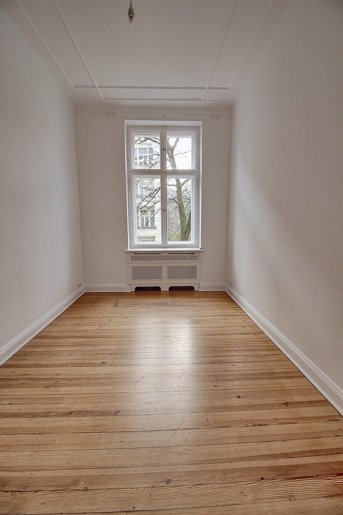 Interior view of an empty, old building room with a floorboard floor, a large window front, and a radiator, from a vanishing point perspective.
