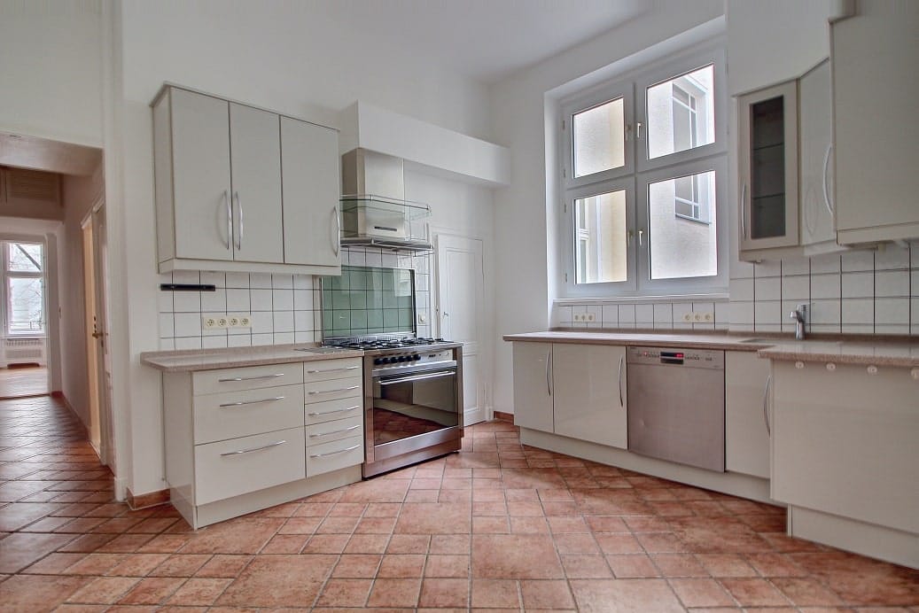 Interior view of an open-plan fitted kitchen with a white bank of units, stainless steel cooker, double window, and terracotta-coloured tiled floor