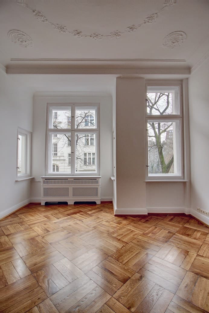 Interior view of an old building flat with herringbone parquet flooring, stucco ceilings and three tall windows with a view of the greenery.