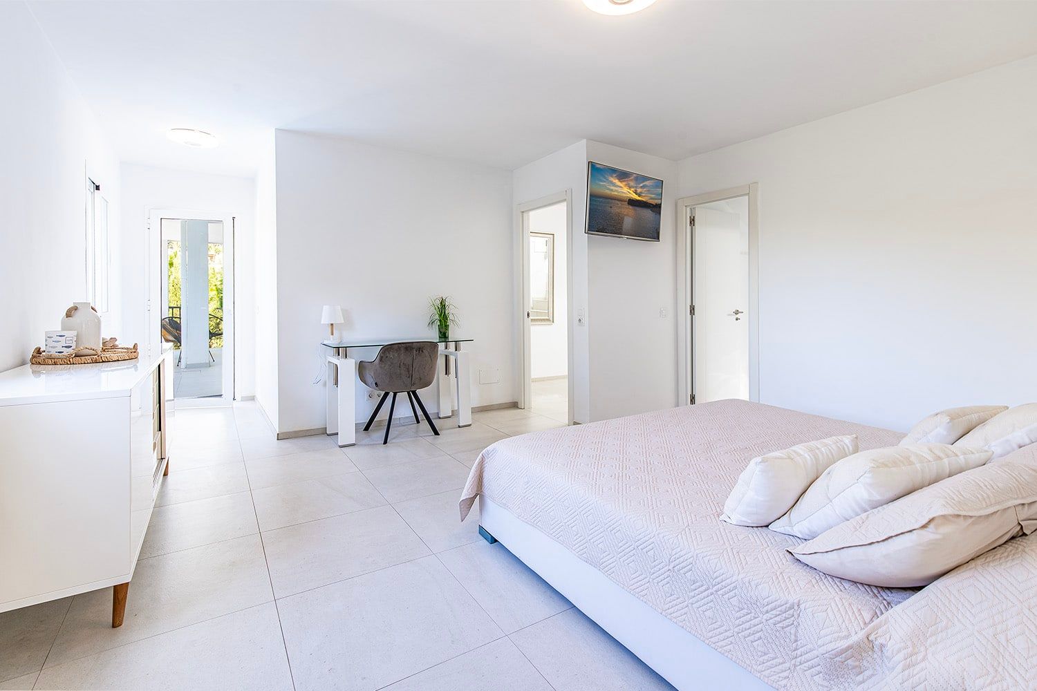 Binnenaanzicht van een lichte slaapkamer met toegang tot het balkon, gezien vanaf het voeteneinde, inclusief bureau en aan de muur bevestigde tv