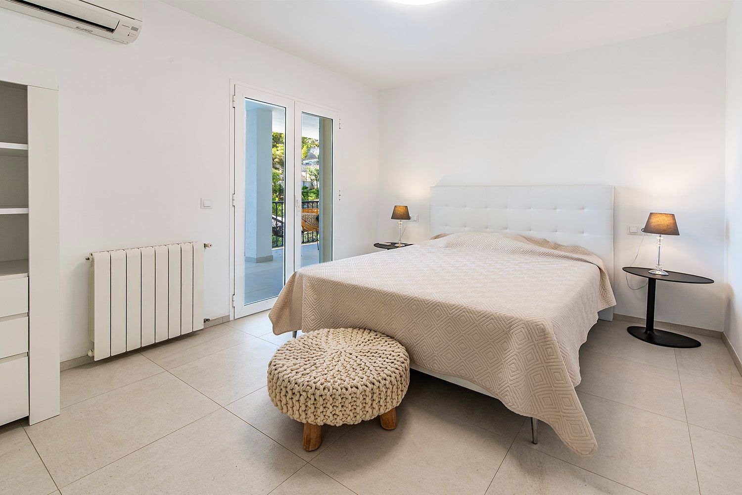Binnenaanzicht van een slaapkamer in een appartement met een tweepersoonsbed, een balkondeur die tot aan de vloer reikt, een tegelvloer en leeslampjes