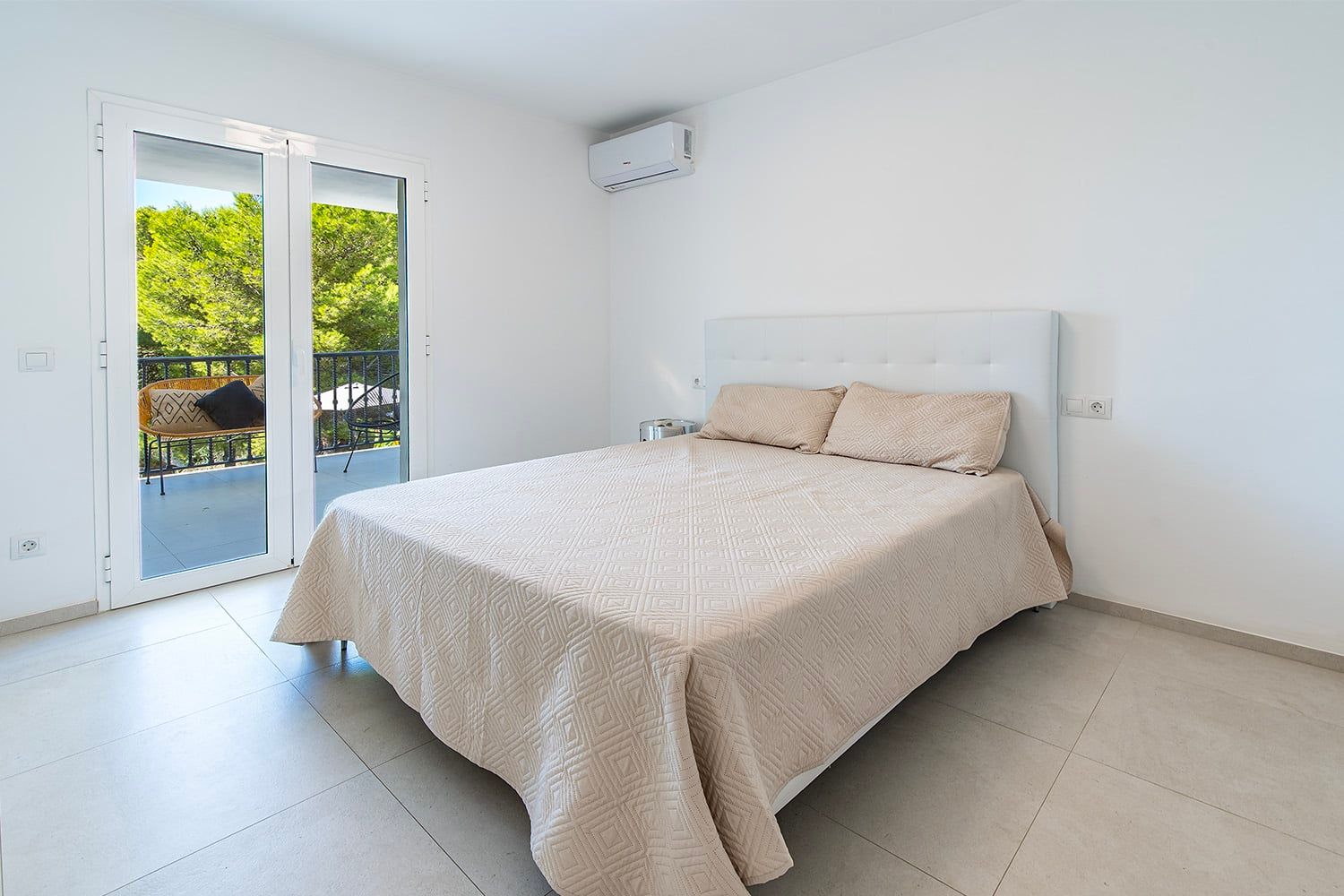 Interieurfoto van een slaapkamer in een appartement met een tweepersoonsbed en een balkon met zitgelegenheid tegen een achtergrond van groene bomen