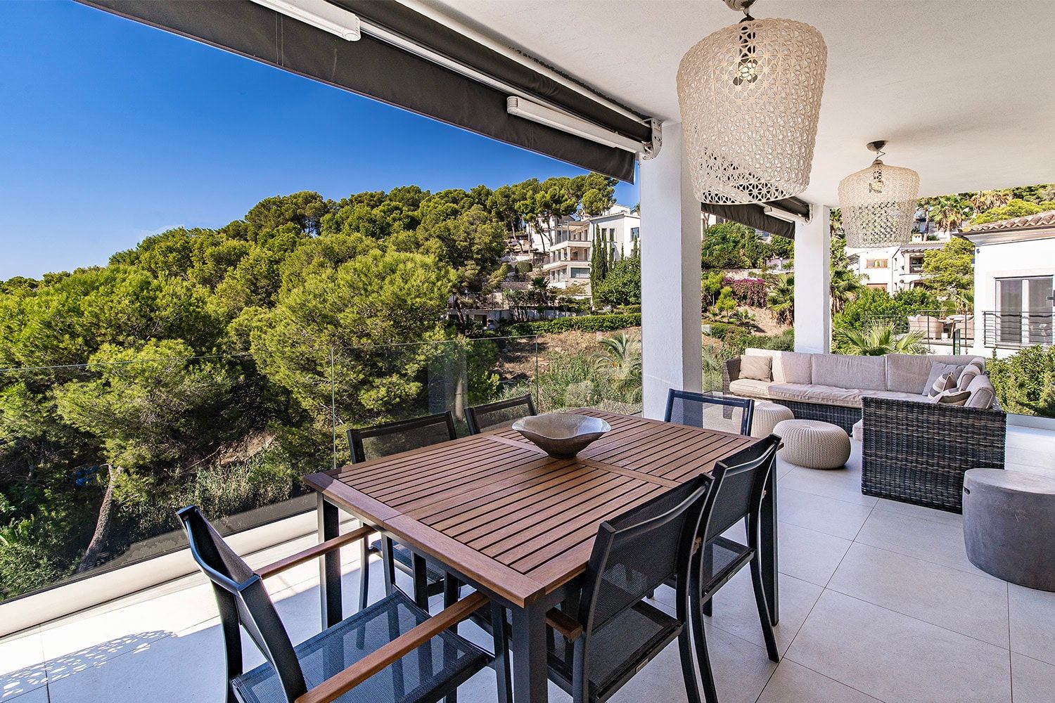 Het terrasgedeelte van een moderne villa met eethoek en loungemeubilair achter een glazen balustrade, met uitzicht op een beboste helling