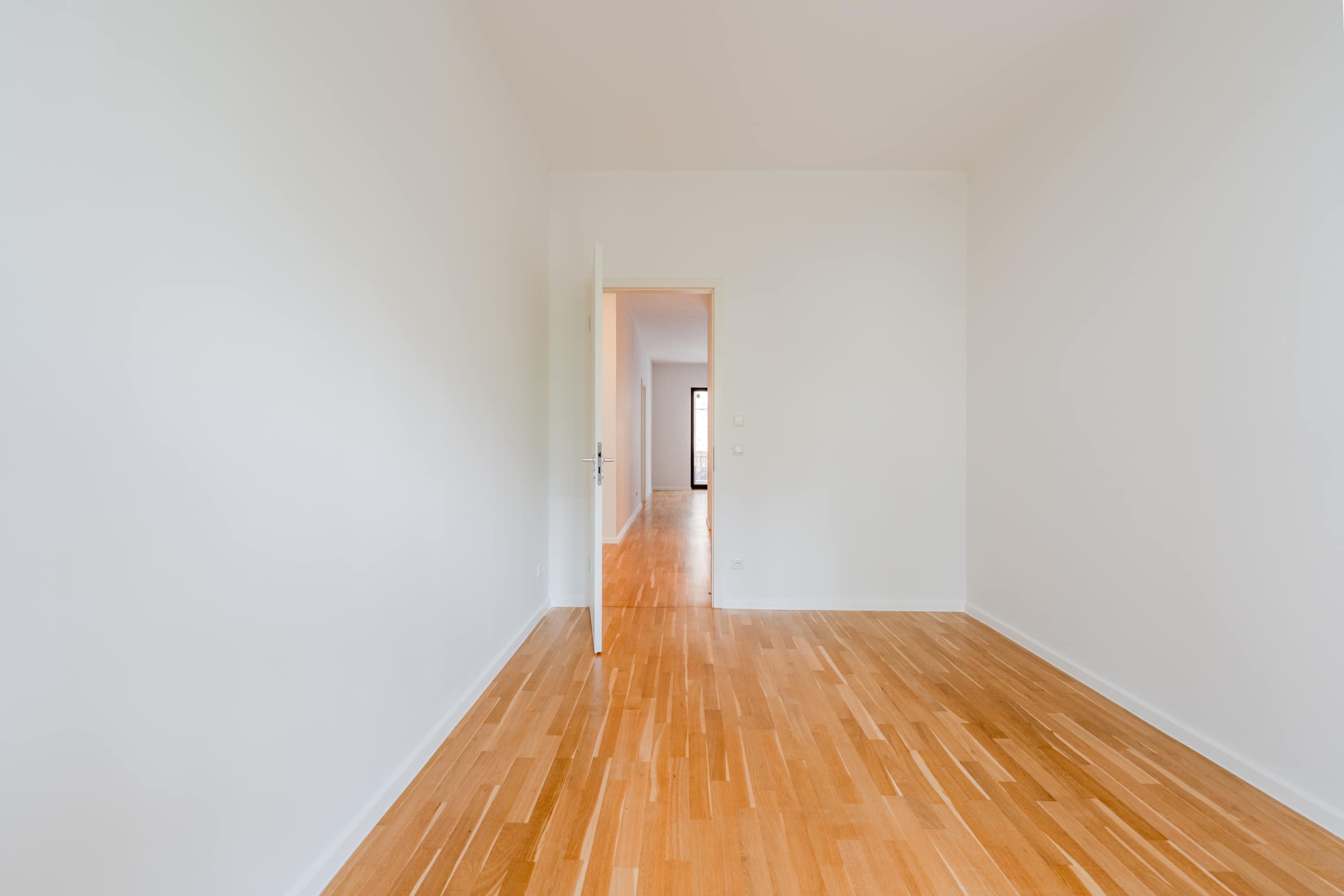 Innenraum eines leeren Apartments mit Parkettboden und weißen Wänden Blick durch geöffnete Zimmertür in den Flur