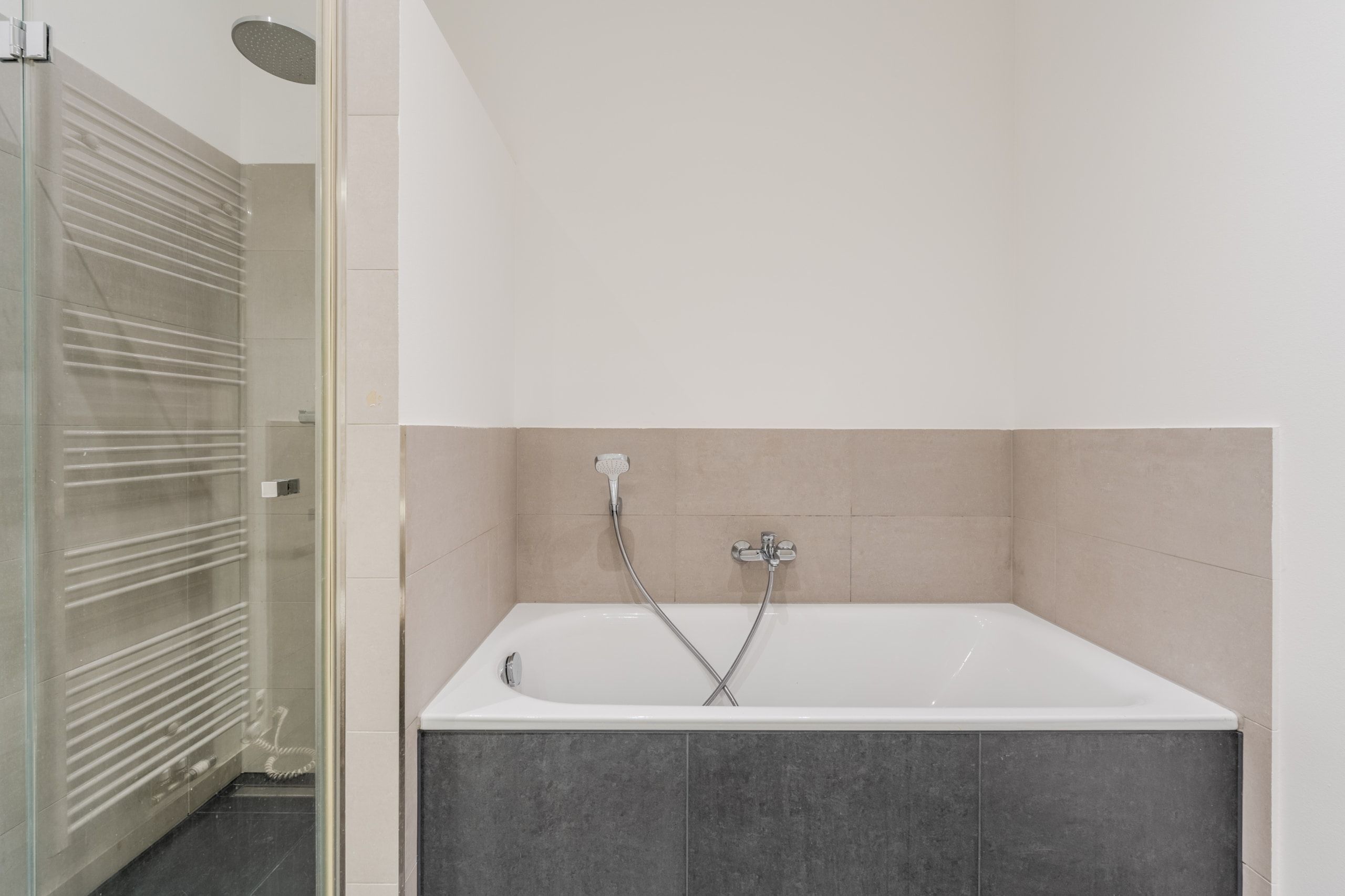 Innenansicht Badezimmer mit eingelassener Badewanne, Handbrause, beige Wandfliesen und Glas-Duschkabine