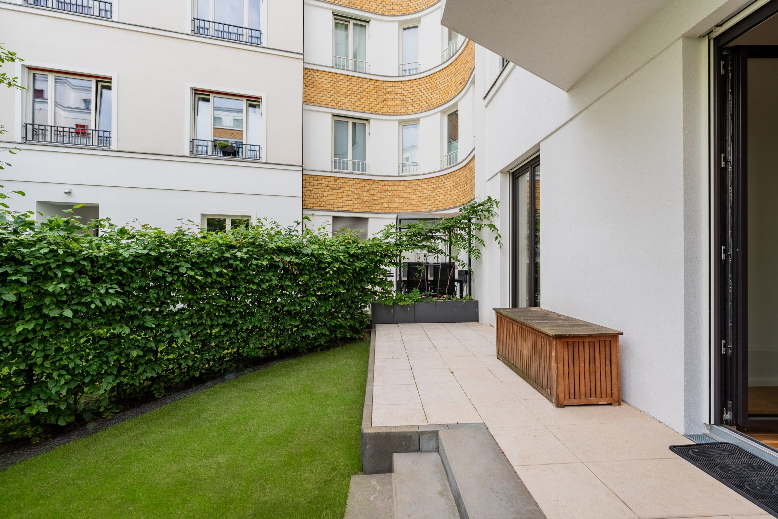 Innenhof eines Mehrfamilienhauses mit Gartenterrasse und Holzbänkchen auf Rasen vor geschwungener Klinkerfassade
