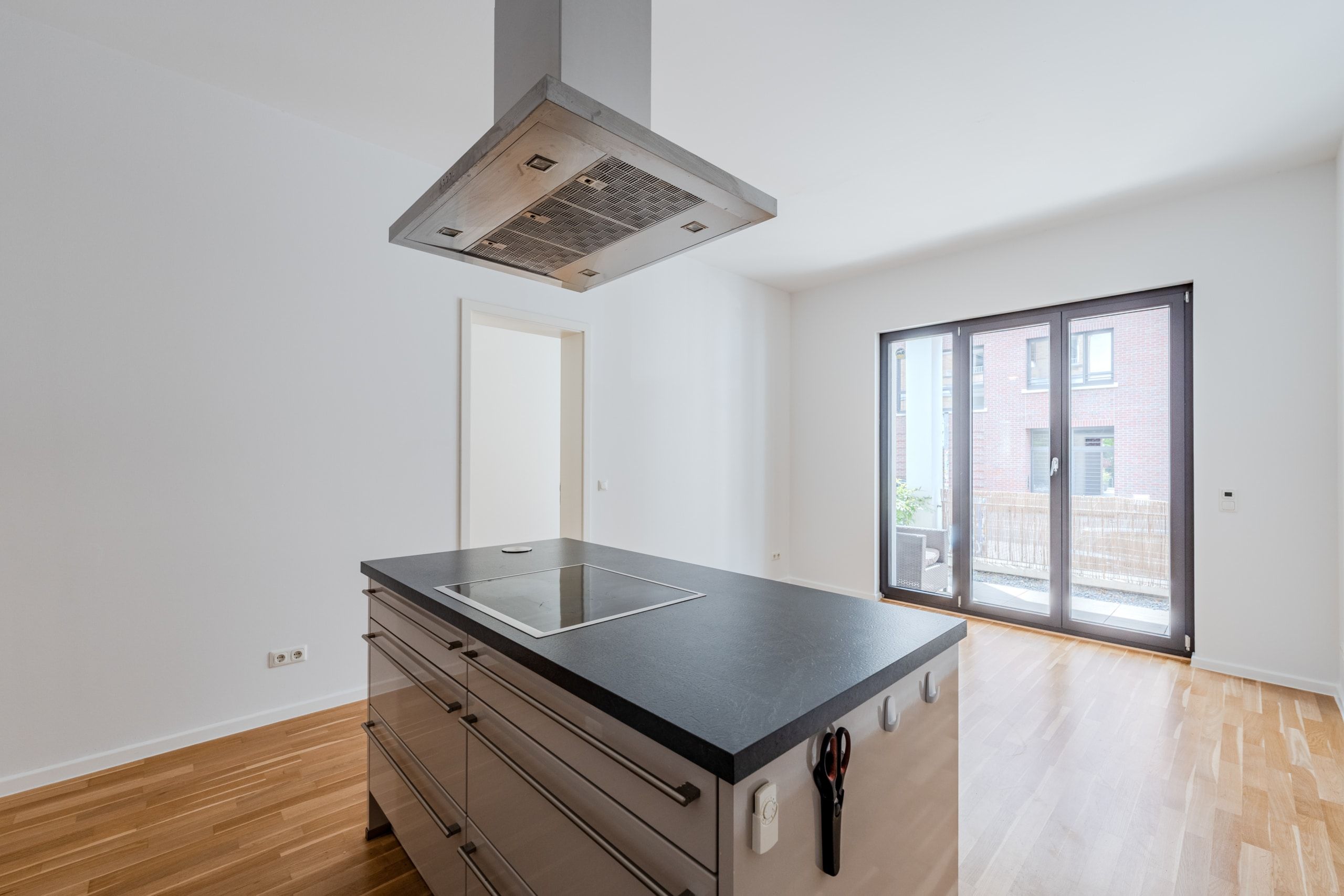 Moderne offene Wohnküche aus Frontperspektive mit Kochinsel, Induktionsfeld, Edelstahlhaube und Balkon vor Klinkerfassade