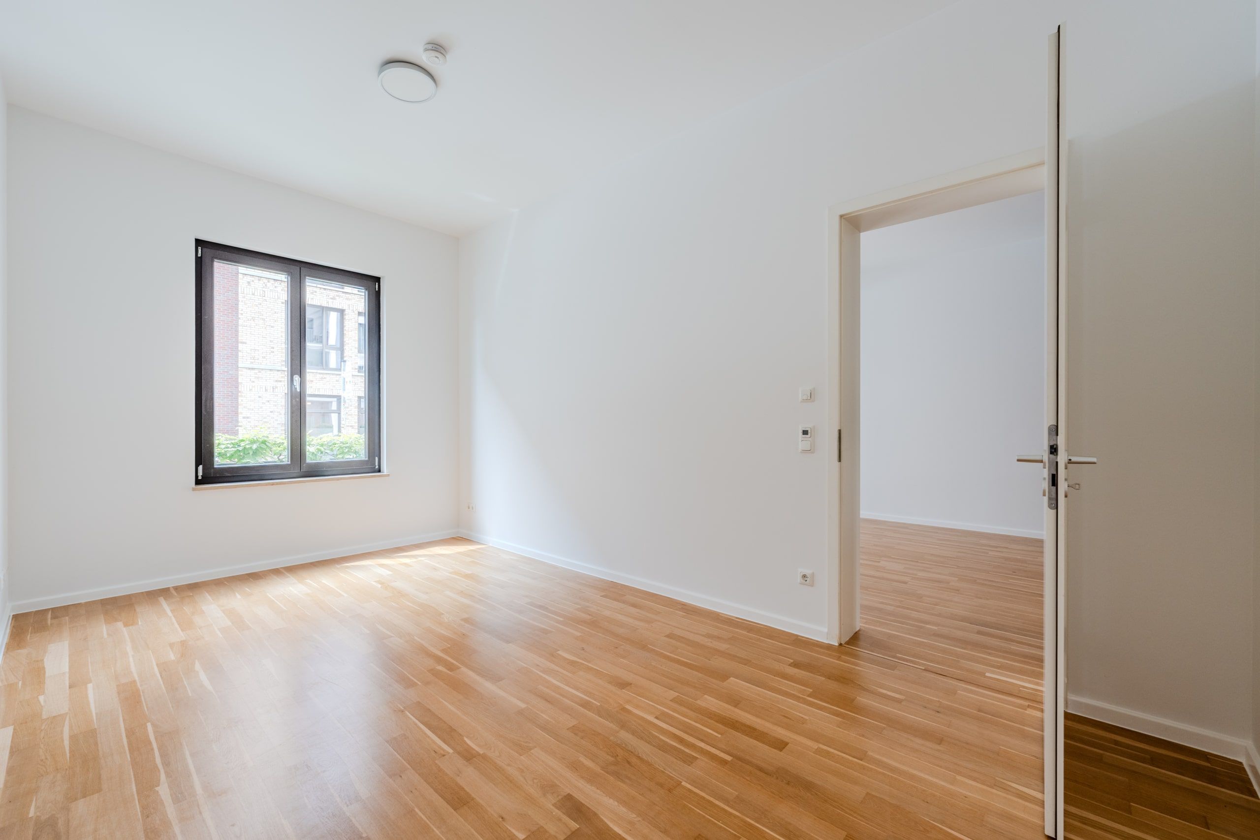 Innenansicht einer Neubauwohnung mit Parkettboden, Fensterfront und offener Zimmertür