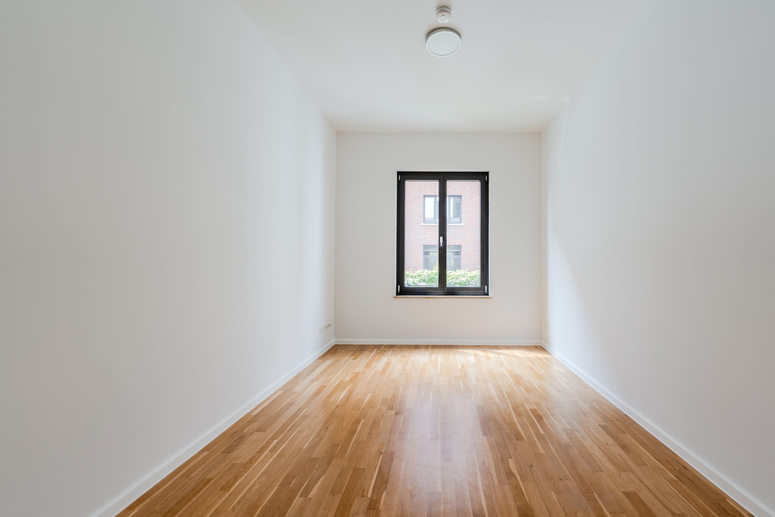 Innenraum eines leeren Wohnzimmers mit Parkettboden weißen Wänden schwarzem Fensterrahmen und Blick auf Klinkerfassade