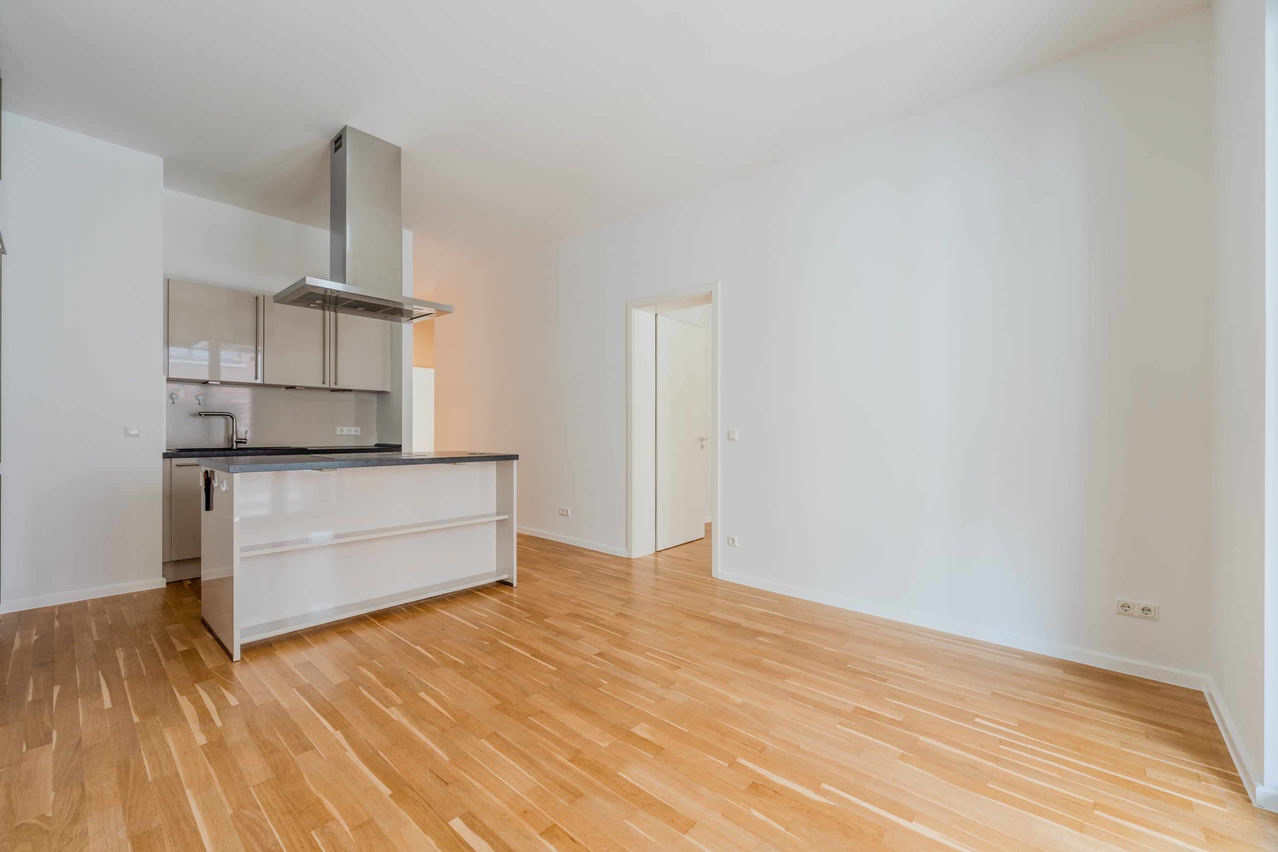 Offener Wohnbereich einer Neubauwohnung mit Kochinsel, Edelstahl-Dunstabzug und Parkettboden aus Eingangsansicht