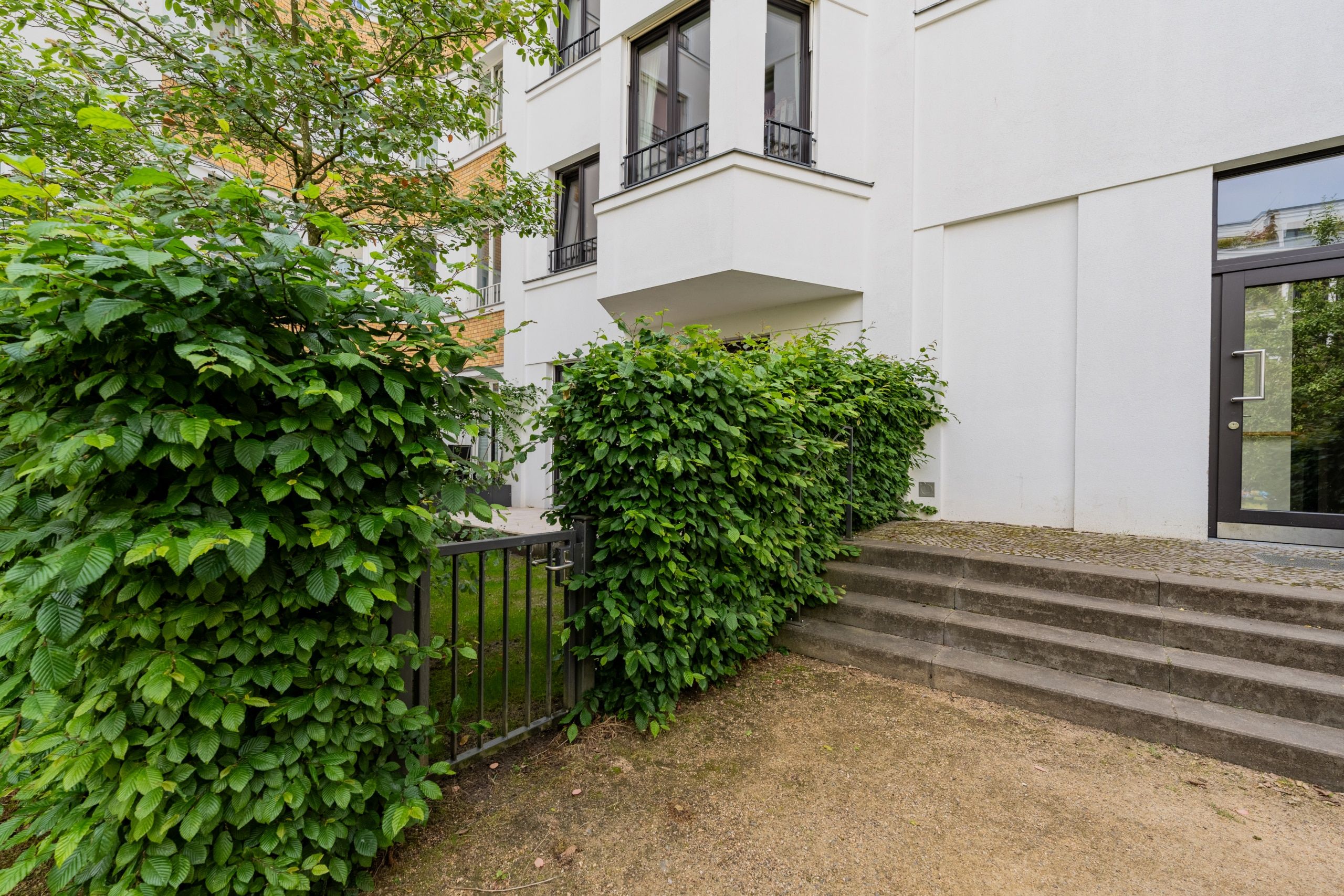 Wohnhaus mit weißer Putzfassade, vorgelagertem Balkon, grünem Heckenzaun, Treppenaufgang und Hauseingangstür