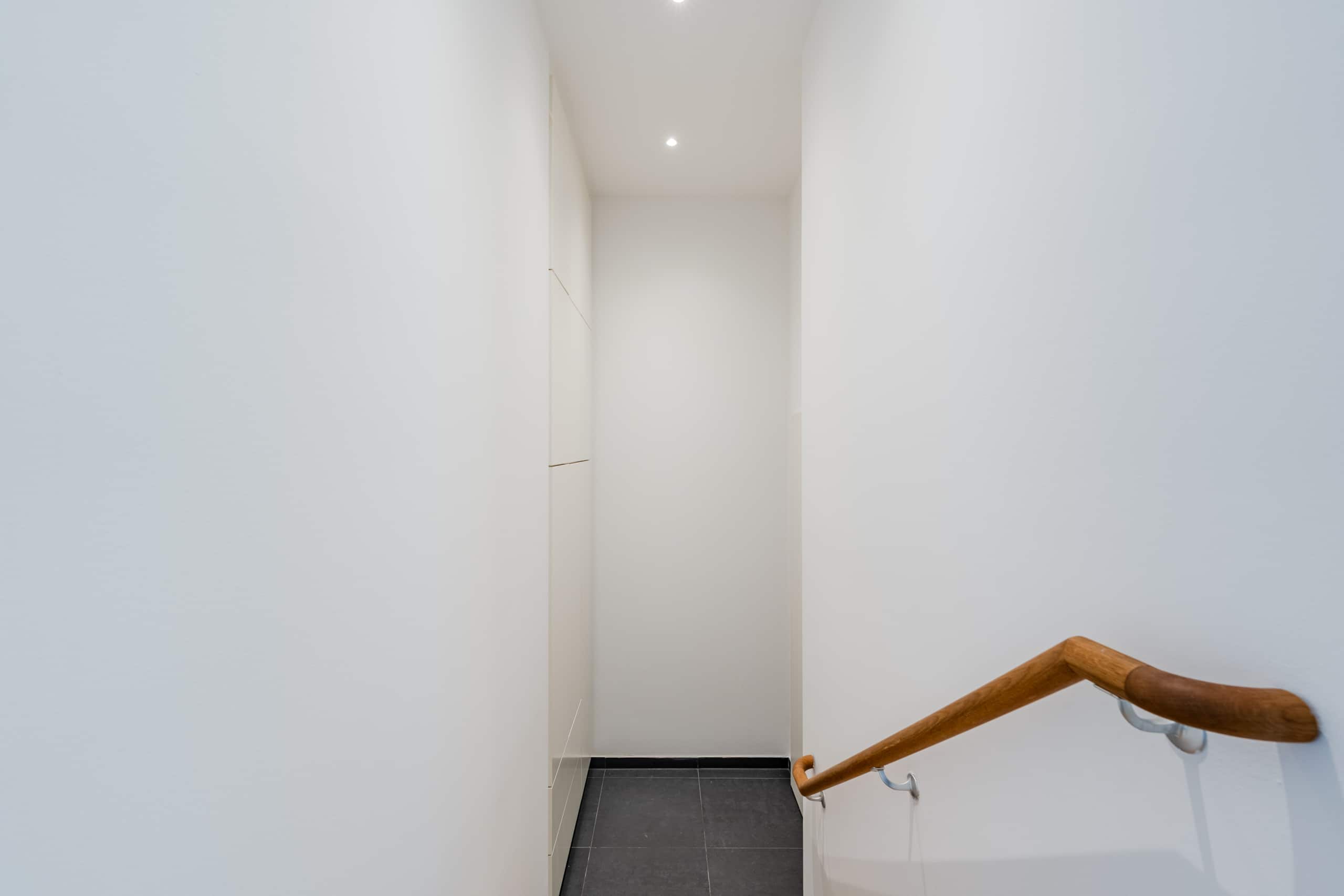Treppenabgang im Wohnhaus mit schmalem weißen Flur, Holzhandlauf rechts und dunkelgrauen Bodenfliesen, Blick von oben