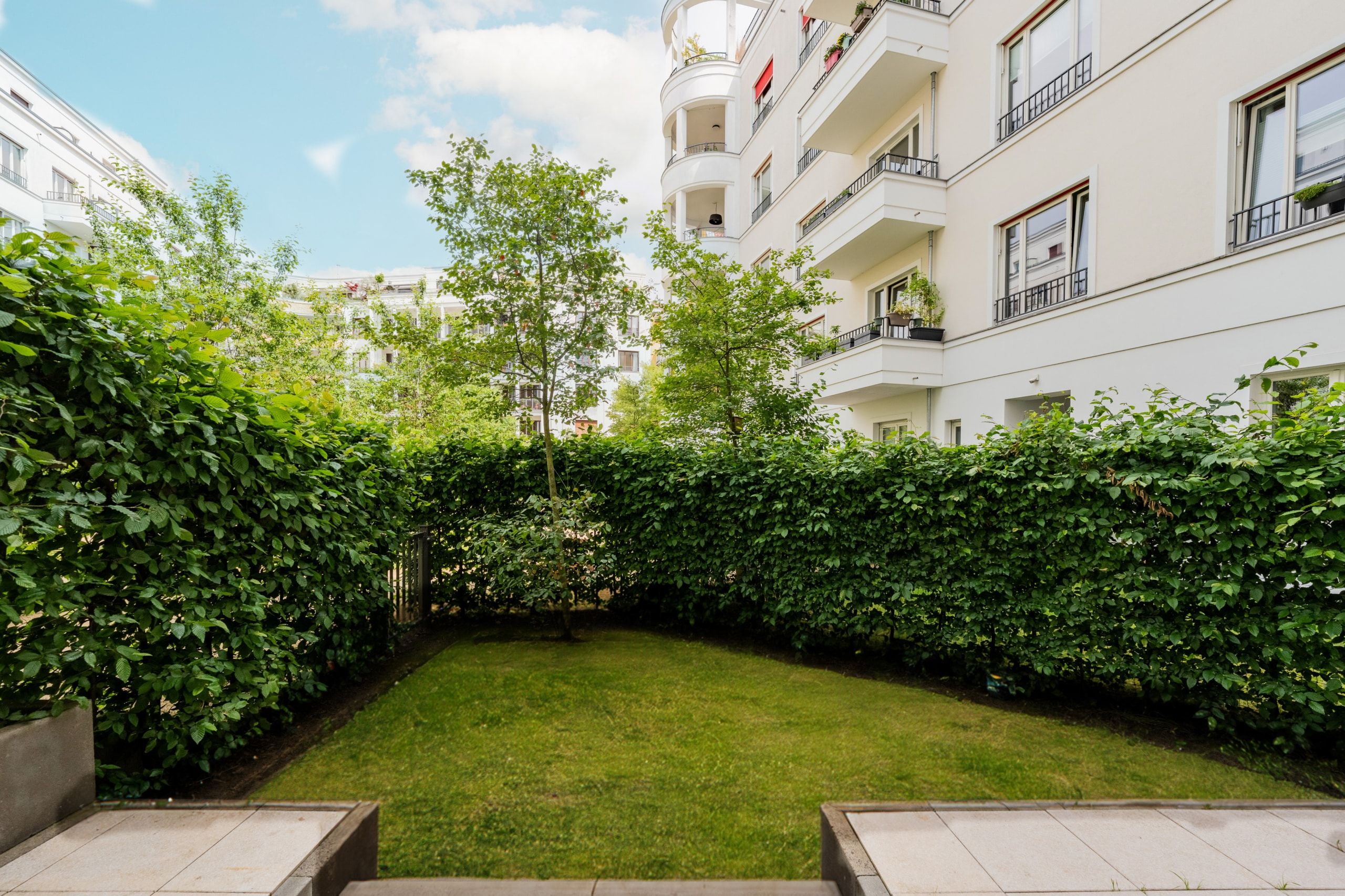 Sicht vom Patio auf kleinen grünen Privatgarten mit blickdichter Hecke vor modernem weißen Mehrfamilienhaus mit Balkonen