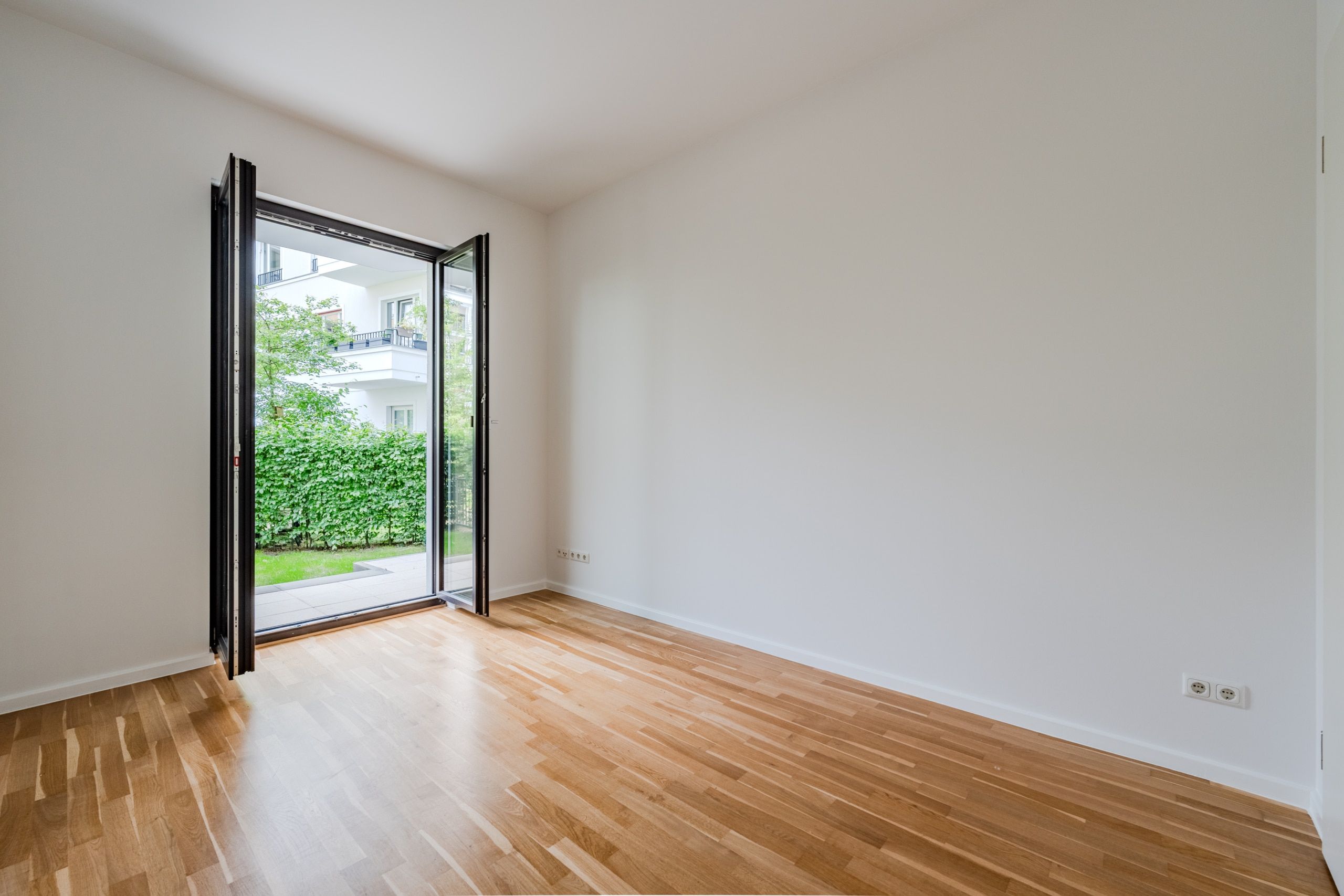 Blick aus leerem Wohnzimmer einer Erdgeschosswohnung mit Parkettboden durch bodentiefe Glastür in grünen Garten