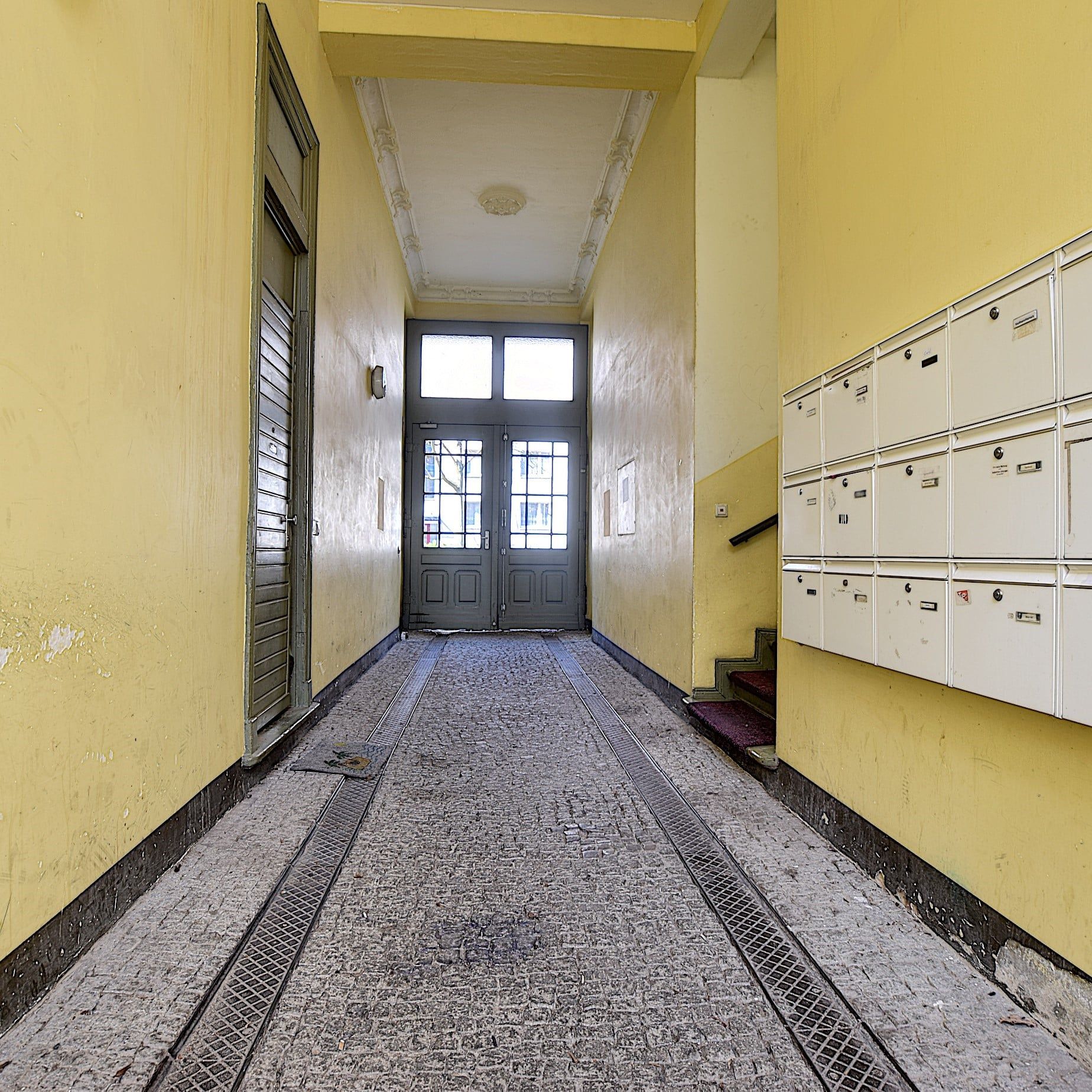 Vista interior del pasillo de un edificio de viviendas con paredes amarillas, suelo de baldosas, puerta de entrada y buzones empotrados en la pared a la derecha