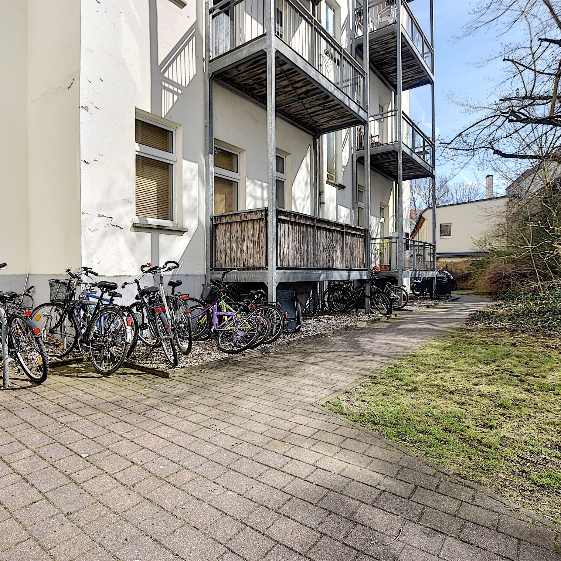 Parte trasera de un edificio de viviendas con tres balcones sobre los aparcamientos para bicicletas del patio interior y un camino empedrado junto a la zona de césped