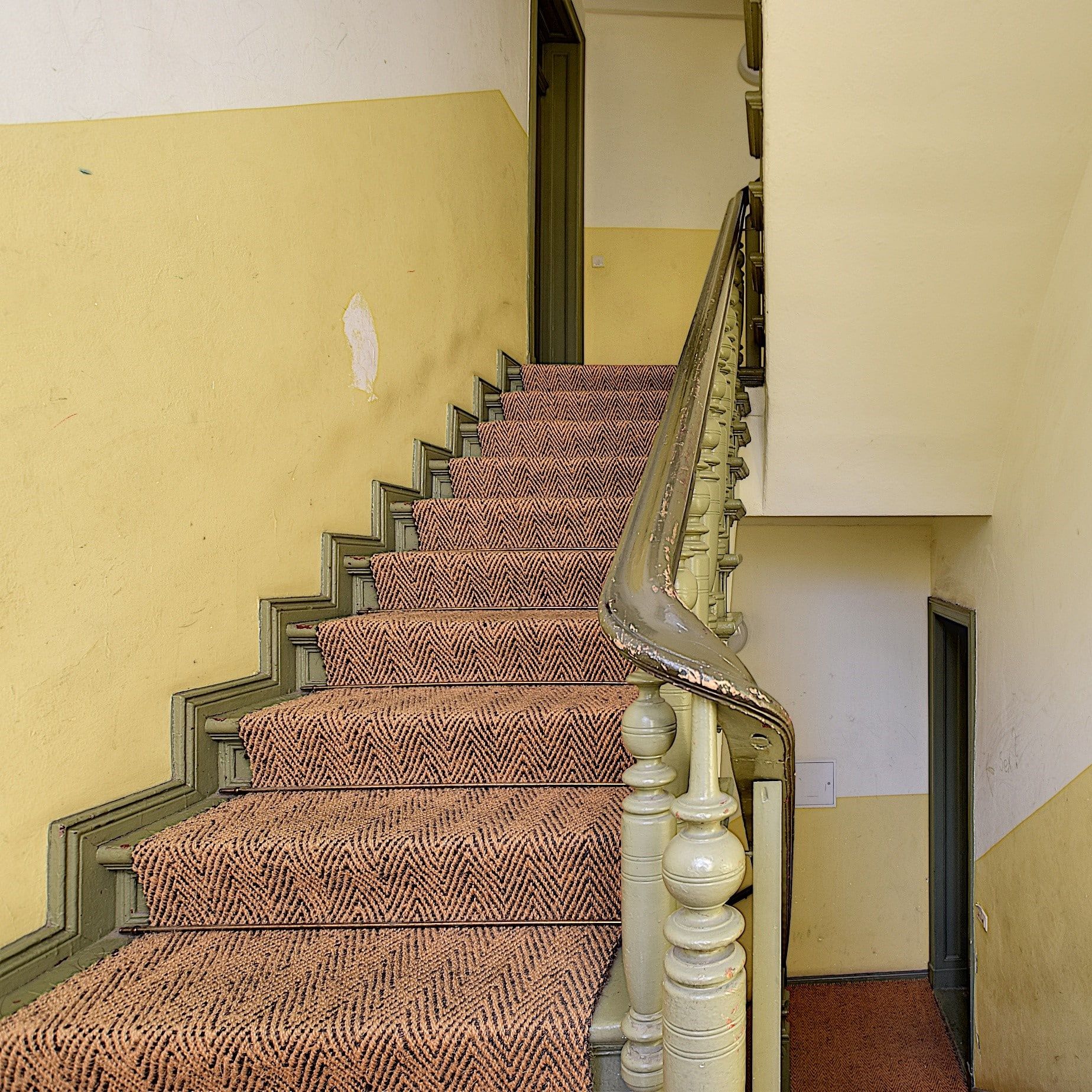 Vista interior de una escalera con un peldaño alfombrado con motivos y una barandilla de madera verde desgastada