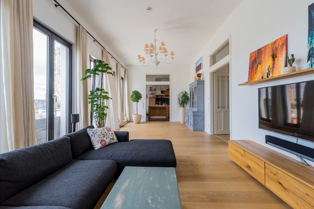 Helles Wohnzimmer einer modernen Stadtwohnung aus Couchperspektive mit bodentiefen Fenstern, Parkettboden und Pflanzen