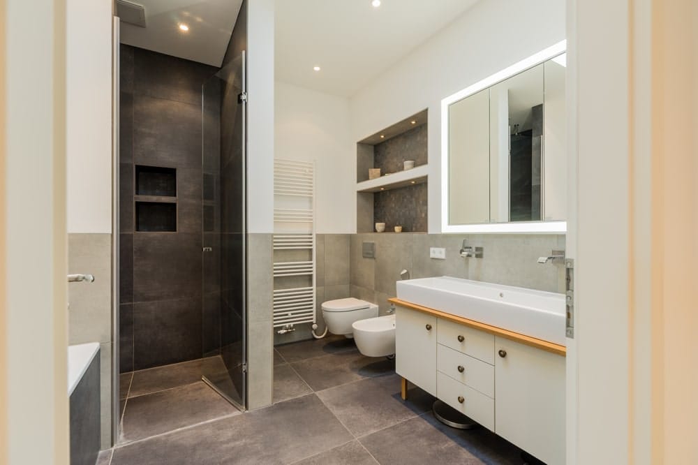 Innenansicht Wohnungsbad mit bodengleicher Dusche Glaswand graue Fliesen Waschtisch mit beleuchtetem Spiegel