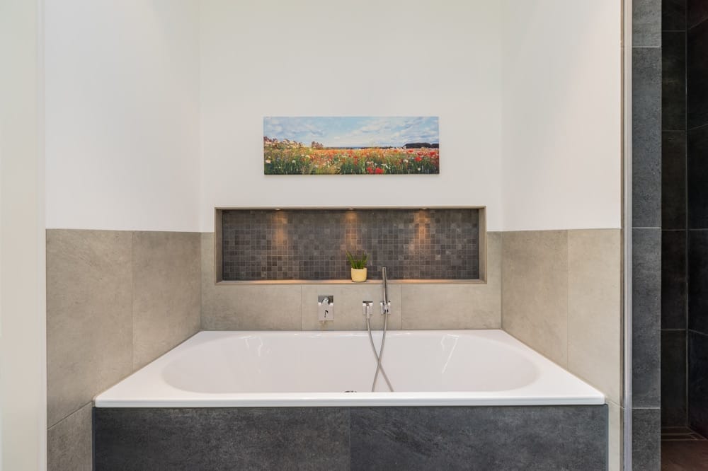 Modernes Bad mit eingelassener Wanne grauen Fliesen beleuchteter Nische mit Pflanze und Blumenbild an weißer Wand