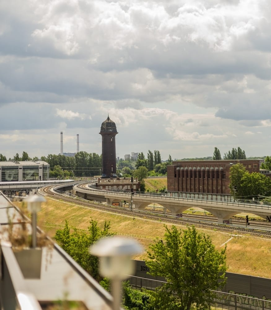 Blick von Dachterrasse auf gekrümmte Bahngleise vor historischem Ziegelturm und Umlandbegrünung