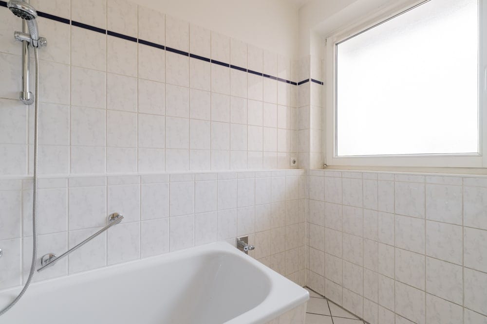 Innenansicht eines gefliesten Badezimmers mit Badewanne, Duscharmatur und großem Milchglasfenster