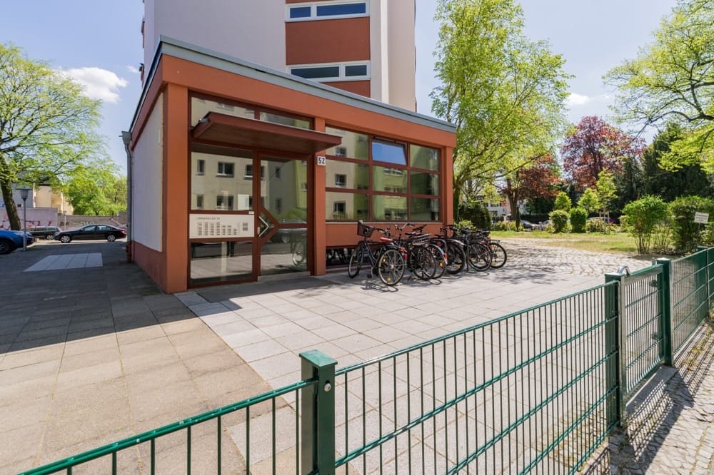 Mehrfamilienhaus mit verglastem Eingangsanbau aus Sicht des Bürgersteigs mit Fahrradständer und umlaufendem Metallzaun