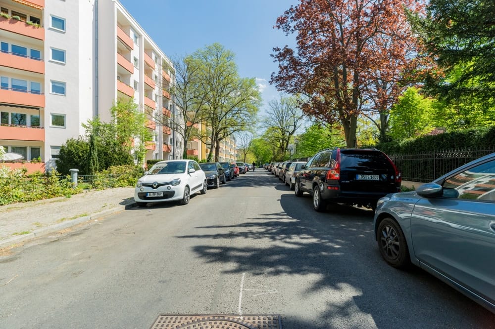 Straßenblick auf mehrgeschossiges Wohnhaus mit Balkonen und parkenden Autos unter Baumbestand