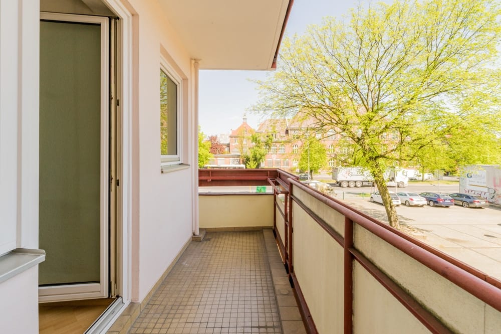 Wohnungsbalkon mit gefliestem Boden und rotem Geländer von innen gesehen mit Blick auf Parkplatz und Baum