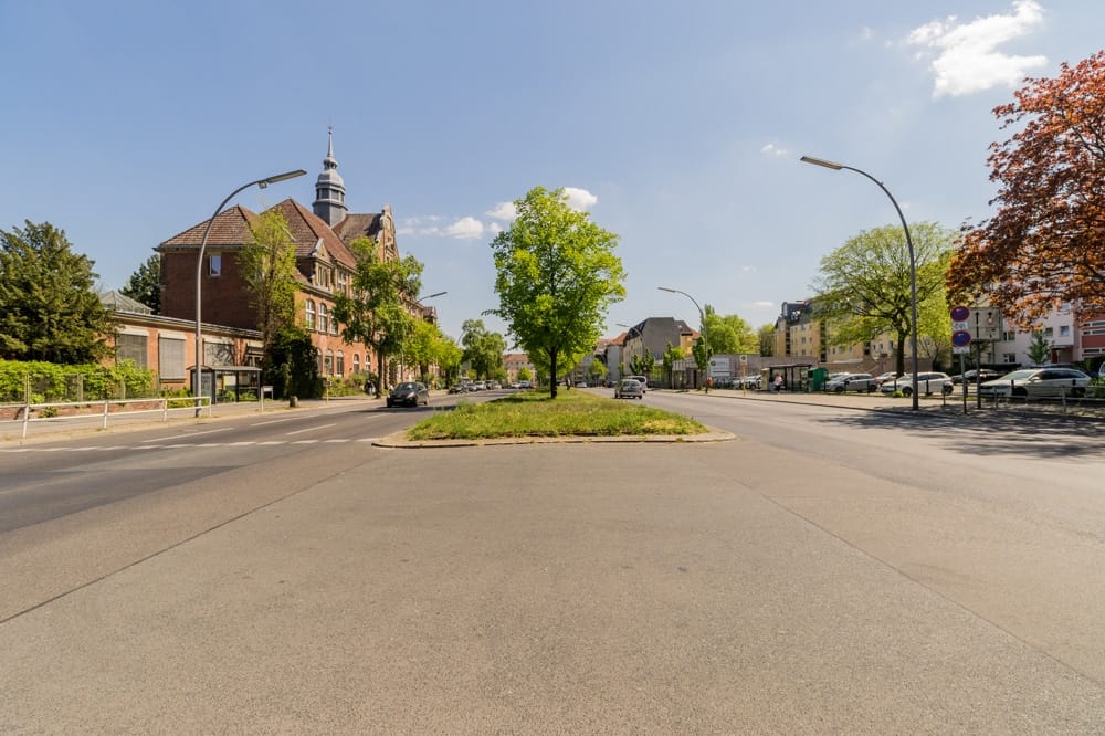 Straßenansicht eines zweigeschossigen Backsteinhauses mit kleinem Turm und begrünten Mittelstreifen