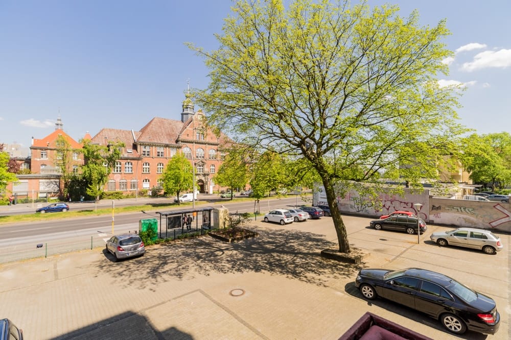 Aussicht von Balkon auf gepflasterten Parkplatz mit großem Baum, Bushaltestelle und rotem Backsteinhaus gegenüber