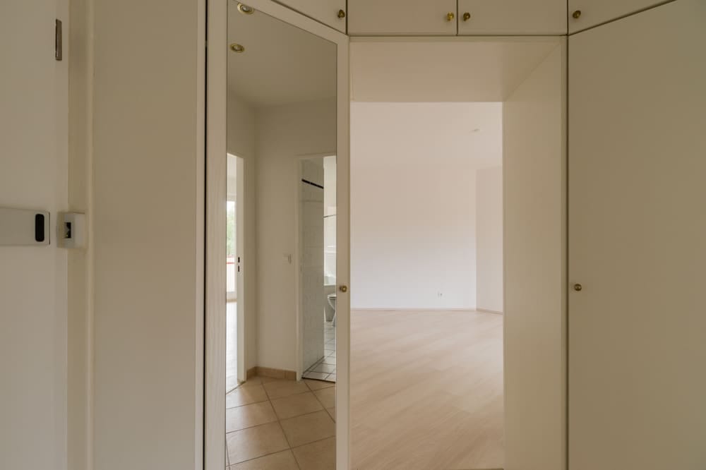 Blick vom Eingangsflur einer leerstehenden Wohnung durch Spiegeltür in den Wohnraum mit hellem Laminat und Badfliesen