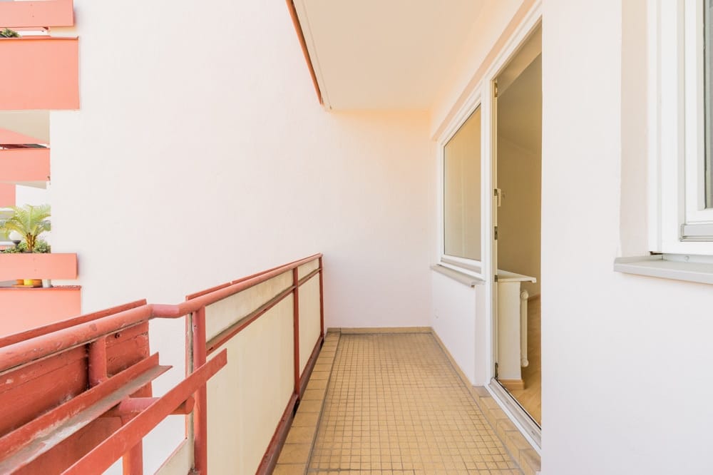 Balkonwohnung mit roter Metallbrüstung, beigem Bodenbelag und weißer Außenwand, Blick durch offene Terrassentür