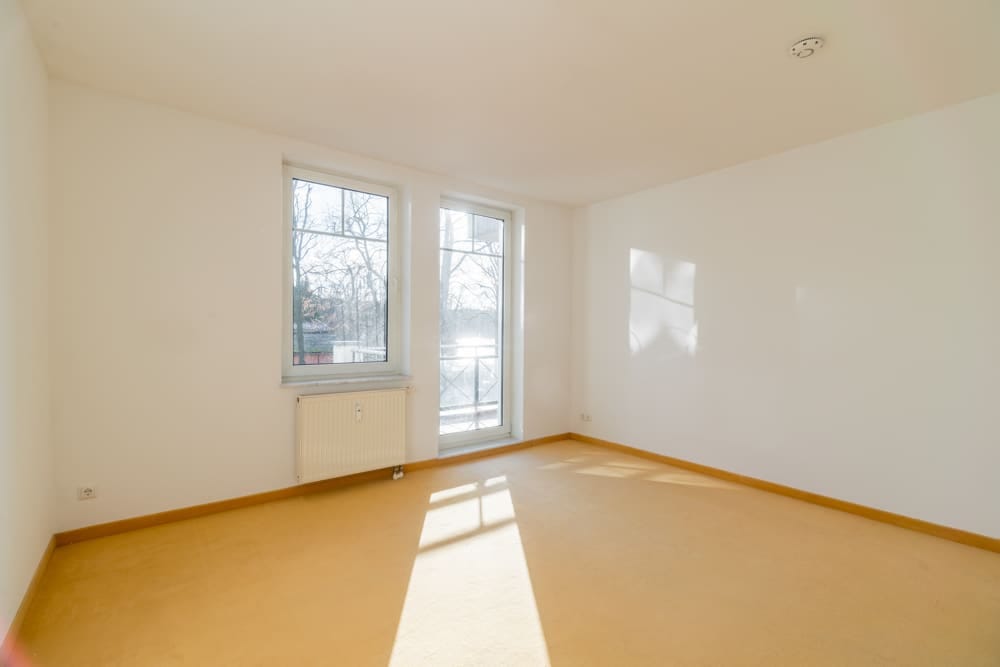 Leeres Wohnzimmer mit bodentiefer Fensterfront und Balkonzugang weiße Wände Heizkörper beiger Teppich