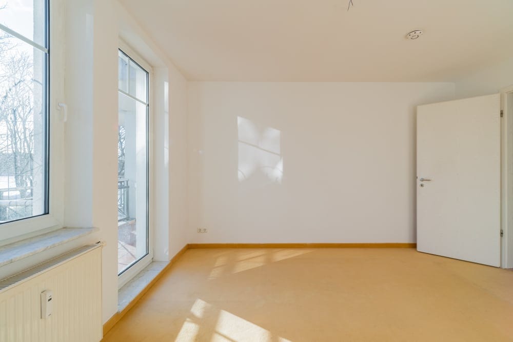 Leeres Apartmentzimmer mit bodentiefer Fenstertür zum Balkon weiße Wände gelber Boden und Heizkörper