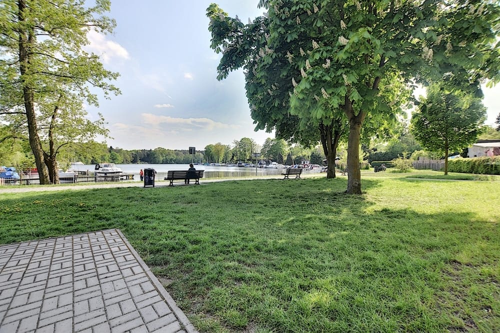 Parkbereich mit Wiese am Seeufer, gepflasterter Weg, Sitzbank unter blühender Kastanie und Bootsanleger