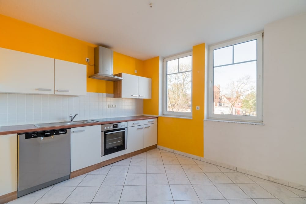 Innenansicht Apartmentküche mit gelber Akzentwand weißen Fronten Fliesenboden und zwei großen Fenstern
