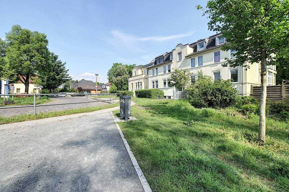 Straßenansicht eines viergeschossigen Mehrfamilienhauses mit weißer Putzfassade, Mansarddach, Vorgarten