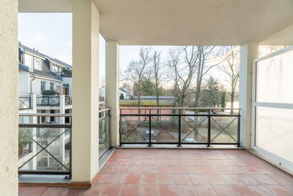 Blick von überdachtem Balkon mit Terrakottafliesen und Gittergeländer auf Wohngebäude und Baumbestand