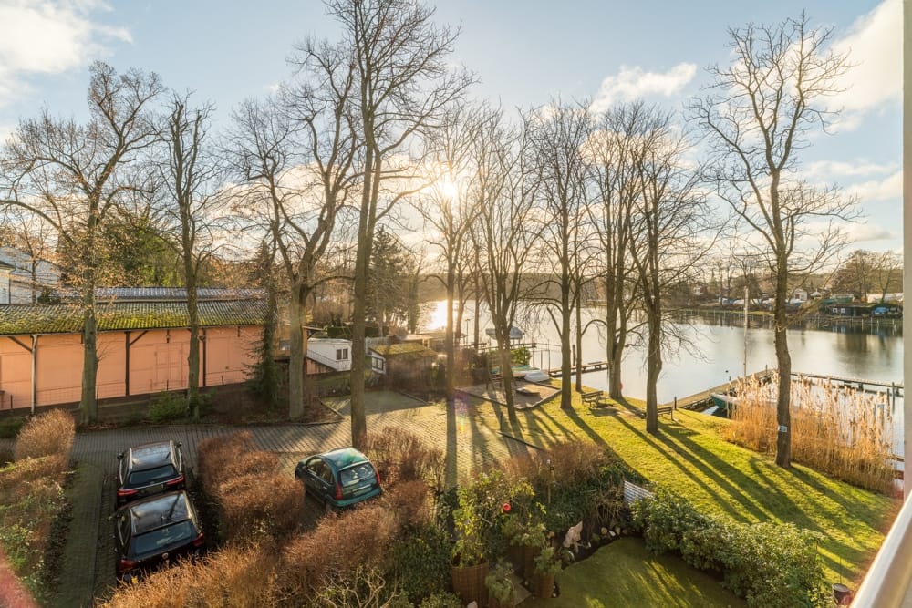 Ausblick von Balkon auf Hauszufahrt mit Parkplatz, Baumbestand und Flussufer mit Bootssteg