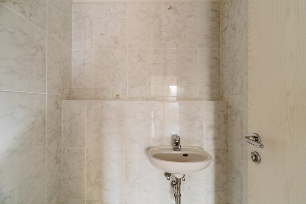 Innenansicht Gäste-WC mit weißem Wandwaschbecken, Marmoroptik-Fliesen und Holztür mit Griff seitlich