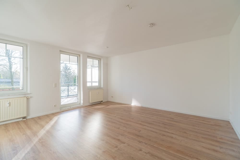 Leeres Apartmentwohnzimmer aus Eckperspektive mit Laminatboden großen Fenstern und Balkonzugang
