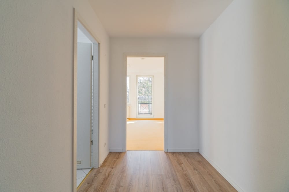 Innenansicht eines Wohnungsflurs mit Holzboden und Blick ins helle Wohnzimmer mit bodentiefen Fenstern und kleinem Balkon