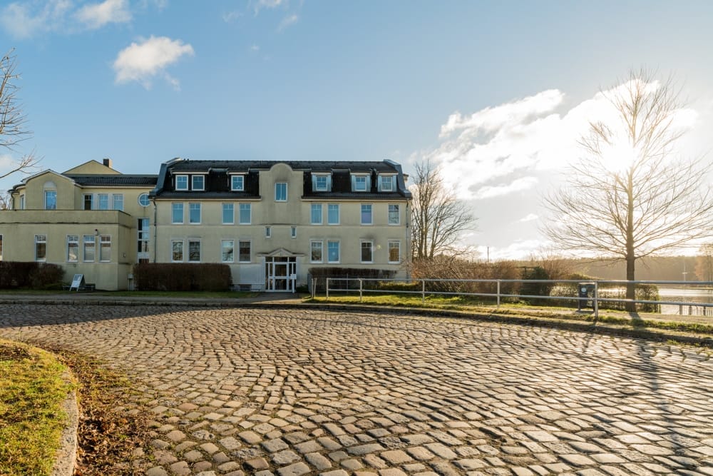 Frontansicht einer dreigeschossigen Villa mit Mansarddach, kopfsteinpflaster und Blick auf Seeufer