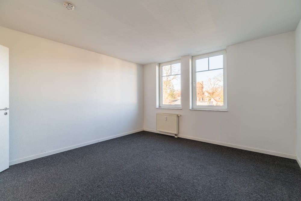 Leeres Zimmer in Wohnung mit grauem Teppichboden, zwei hohen Fenstern und weißem Heizkörper unter den Fenstern