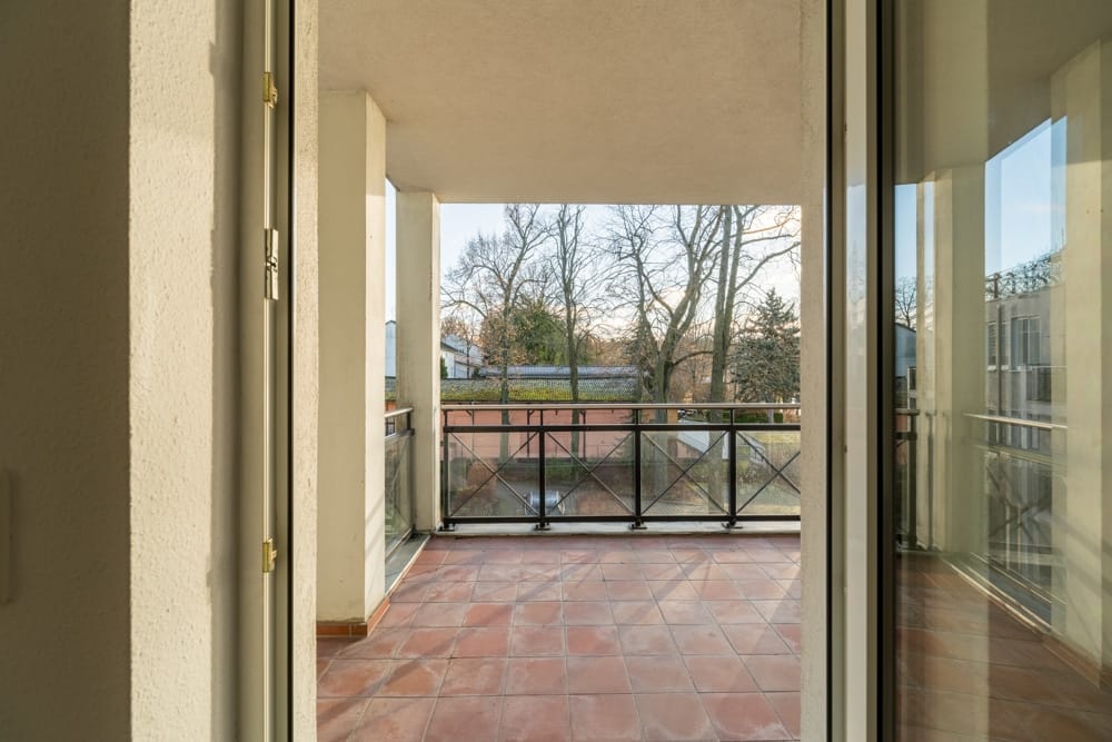 Blick durch geöffnete Balkontür auf Wohnungsbalkon mit terrakottafarbenen Fliesen und Metallgeländer Richtung Garten und Bäume