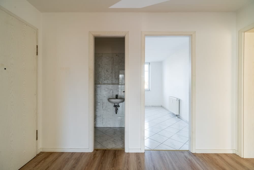 Innenflur einer Wohnung mit Laminatboden, Blick in Badezimmer mit Waschbecken und helles Zimmer mit Fliesenboden und Fenster
