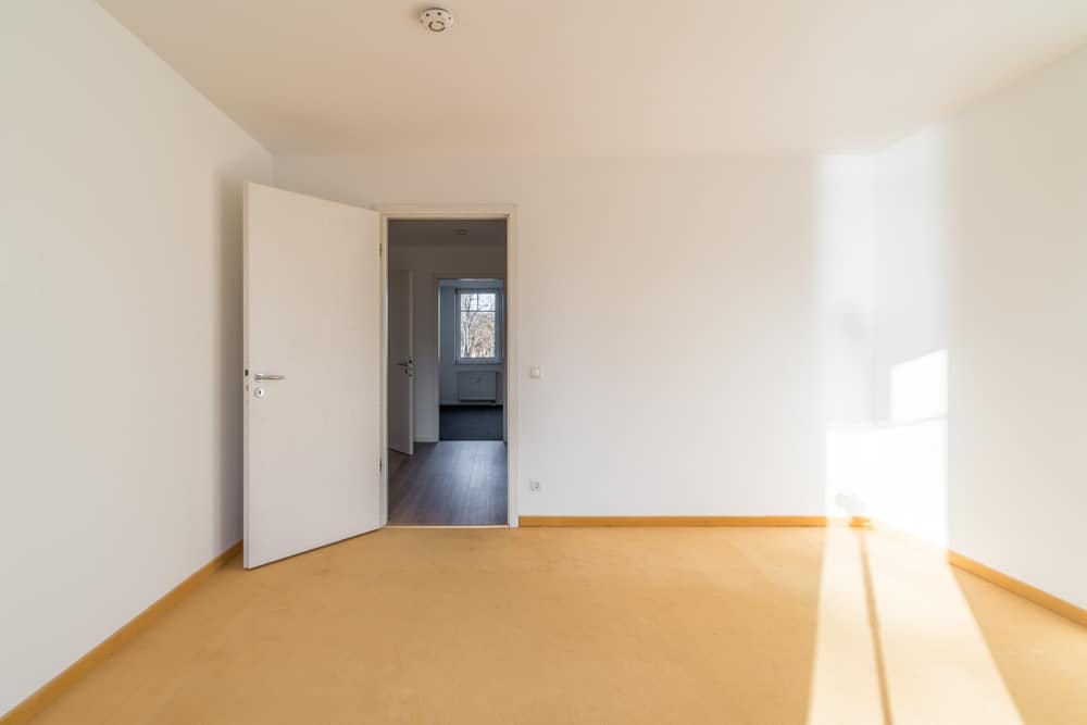 Innenansicht leeres Zimmer mit weißem Anstrich, offener Tür zum Nebenraum mit Fenster und sonnigem gelblichen Boden